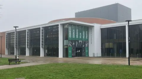Amy Holmes/BBC A white block building of long windows with green banners reading 'comedy, dance, drama, film, music and panto" - this is the Grove Theatre in Dunstable 
