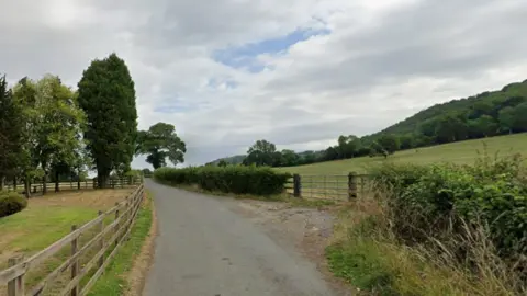 A narrow country lane surrounded by open countryside in Gloucestershire. The lane is bordered by post and rail fencing to the left, and hedgerow with a metal fence leading to a sloping field on the right. There are trees dotted around, and there is woodland on the hill in the distance.
