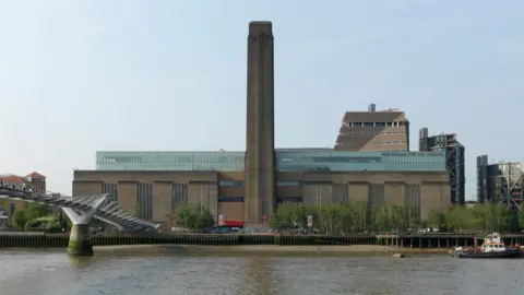 Tate Photography The exterior of London's Tate Modern gallery, viewed from across the river. The River Thames is visible in the foreground at the bottom of the picture, with a grey pedestrian bridge spanning the river in the bottom left corner. A large brown building is situated on the river bank, with a top floor made of glass. A large brown tower rises from the front-middle of the building, by the riverside. There is a row of green leafy trees along the river bank. The sky is pale blue and sunny.