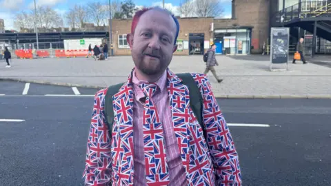 Julita Waleskiewicz/BBC Bartly is dressed in a salmon pink shirt with white stripes, and a british flag tie and jacket. He has dyed pink/purple hair and a brown goatee beard. He has rucksack straps over both shoulders and is standing with the station in the background.