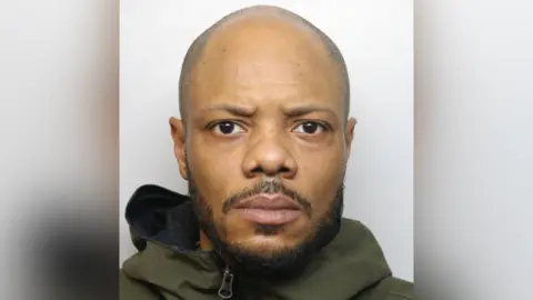 Sam Layne mugshot. He is bald and is wearing a green hooded coat. He has a short beard.