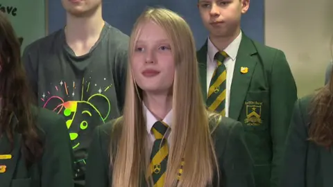 A girl with long blonde hair, wearing a green school blazer with a green and yellow striped tie.  Two boys are standing behind her