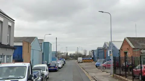 A Google Streetview image of Strickland Street in Hull. The road is lined by industrial looking buildings which have cars and vans parket outside. A footbridge is visible in the distance. 