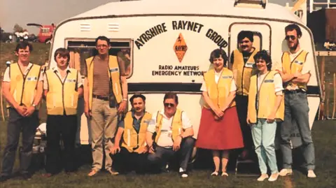 Raynet An old eighties image of the Ayrshire Raynet volunteers, standing in front of their mobile office - a caravan with the Raynet logo on the side. It appears to have been taken at some kind of public event.
