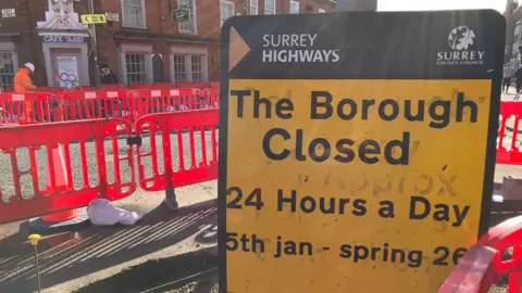 A yellow and red road sign announcing that The Borough in Farnham is closed from January to Spring sits in front of a row of red plastic road barriers around a set of roadworks.