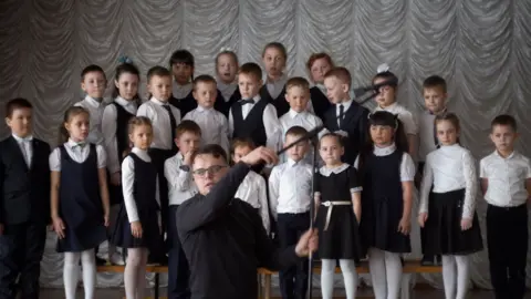 Pavel Talankin School choir -- girls in white tops and black dress or skirt, boys in white shirt and trousers, one boy with black jacket, one boy in waistcoat and bow tie
Pavel Talankin in the bottom centre in black setting up microphone
