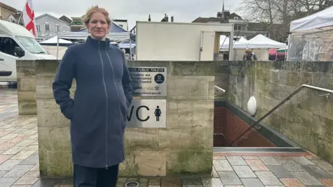 Councillor Victoria Charleston standing by the Market Square toilets in Salisbury. She has blonde hair which is tied up and is wearing a long navy blue coat. Her hands are in her pockets and she is looking directly at the camera and smiling. 