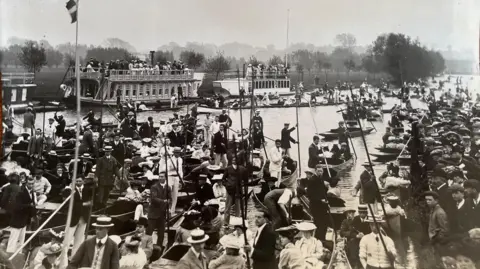 Sarah Hosking’s archive A black and white archive image of people on boats at the river in Oxford and three college barges lined by the bank.