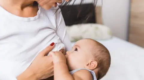 A mother breastfeeding her child. The mother is wearing a white long sleeve stop and sitting on a bed.