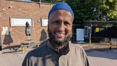 Jama Hussein, who has a black beard, is wearing brown clothes and a blue hat. He is stood outside a social club in Barton Hill