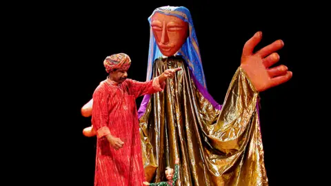 Serendipity Arts A performer in a red tunic and patterned head wrap manipulates a small puppet on a stage beside a towering figure draped in gold fabric, with an oversized painted head and large outstretched hands against a black background.