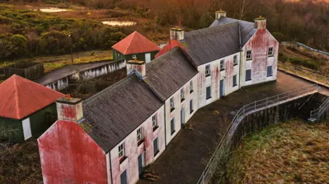 Jay Curtis Close up drone shot of a street in the village. The houses are white with black bricked roofs. They are derelict with red marks on their sides.