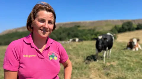 Margaret Lees Junior is standing in a field with cows in the background. She is wearing a pink polo shirt.