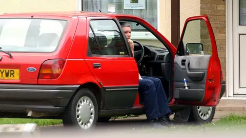 PA Media Huntley is half-sitting in a red Fiesta car, parked outside a house with a white front door. He is wearing dark blue jeans or trousers. He is sitting sideways in the driver's seat. The driver's side door is open and he has his feet on the ground.
