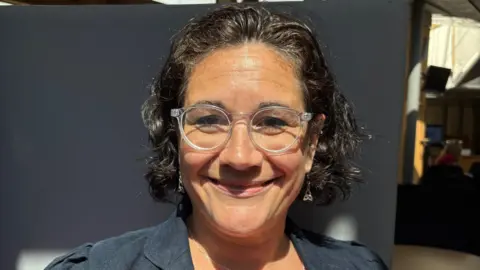 Katie Hagmann wears clear glasses and a dark blazer. She has short dark hair and smiles at the camera