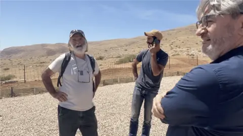 A man in a white t-shirt and a man in a dark coloured t-shirt stand next to eachother. One is on the phone.