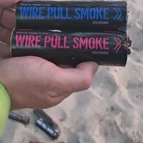 Swanage Coastguard Team Hands, with the cuff of a high-vis jacket visible, holding tubes of smoke bombs, with a number of discarded tubes on the sand below.