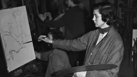 Getty Images A black and white picture of Susan Collinson, a woman with short dark hair and wearing painting overalls, while she paints a picture.