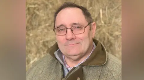 Farmer Steve Glover looks at the camera. He is wearing a green and brown tweed jacket and in the background there is a large pile of hay.