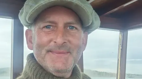 Jeremy Gower grins at the camera in the wheelhouse of his boat. He is wearing a large grey woollen cap and a khaki green roll-neck jumper.