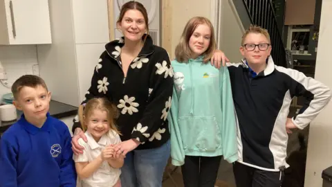 A woman standing inside her house with four young children all lined up alongside her.