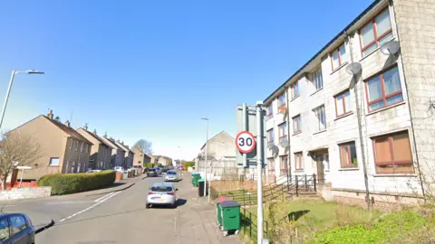 Terraced housing and flats line the street. There are two large green bins. It is a sunny day and the sky is blue. There are cars parked along the street.
