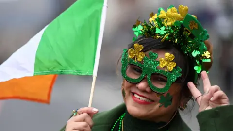 A woman dressed in green, with an Irish flag in her hand. She is wearing Shamrock glittery glasses and a headband. She also has Shamrocks painted on her face.