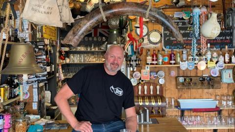 Man sat on a bar surrounded by a tusk and other items such as a bell