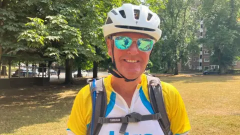 BBC/Seb Cheer A man wears a bright yellow cycling jersey and white helmet. He is standing in a grass-covered park.