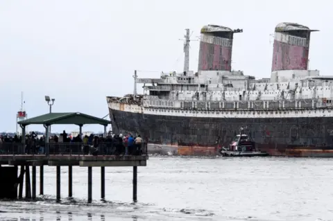 Matthew Hatcher/Reuters Onlookers watch the SS United States as she is towed out to sea by tugs. The ship looks old and rusty.
