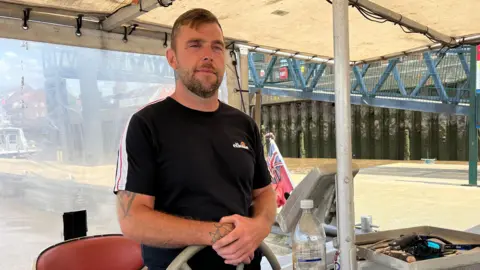 BBC Ben Ellis standing behind the ferry's helm. He is resting both hands on the top of the wheel. He has short brown hair and is wearing a black t-shirt with a white and red stripe down the sleeve. It is a sunny day. He is under an off-white awning.