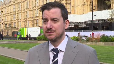 Mike Reader, who has dark hair, a short moustache and beard, is wearing a grey suit with a white collared shirt and blue and white tie. He is standing on a green outside the Houses of Parliament in London on a bright and sunny spring day. 