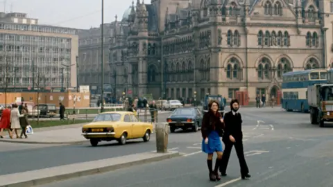 A historic image of Bradford in the 1970s with a couple crossing a busy road outside City Hall