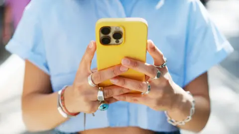 A woman wearing a blue crop top and jeans holds a mobile phone in a yellow case as she looks down at the screen, her face cropped out of the picture.