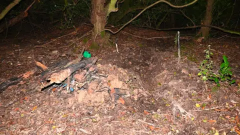 Devon and Cornwall Police Image taken at night with some kind of artificial lighting or flash lighting it. Shows two tree trunks in the background and a forest floor. Towards the middle of the image is a pile of logs and other material. A green petrol can is also visible and what appears to be an indentation in the ground to the right of the pile of logs.