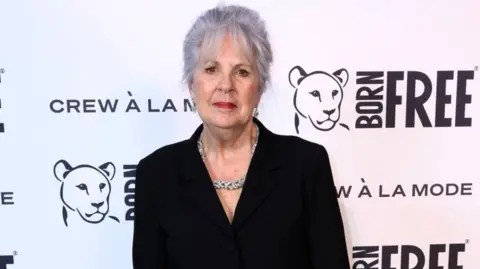 PA Media A woman with short hair, a large necklace and wearing a black suit, stands in front of a white board with the Born Free logo.