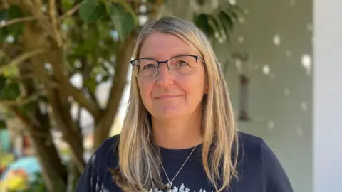 Sheena Brockie is standing in her garden, with a tree behind her. She is wearing glasses and a navy t-shirt and is looking into the camera.