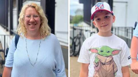 On the left is Lucy - a woman with blonde hair and glasses. She is wearing a light blue jumper and blue necklace. On the right is her young son Toby, wearing a red, white and blue hat and a white T-shirt with the Star Wars character Grogu on it.