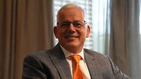 Ann Gannon / BBC A head and shoulders image shows Michael Hales smiling into the camera. He is sat in a room wearing a grey suit with orange trim and a bright orange tie.