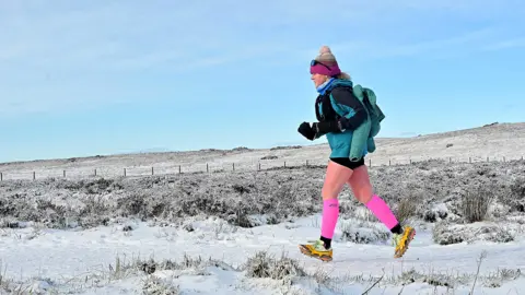 A woman jogging along a snow covered path with fields and a fence in the background also covered in snow. She is wearing yellow trainers with spikes, bright pink socks, a blue top, a pink bobble hat and sunglasses up on her head. Above he blue skies