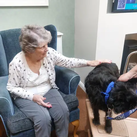 Barchester Healthcare An elderly woman sits in an armchair in a care home and strokes a small black dog that is stood on a table so she can reach it.