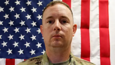 US Army Portrait of Jeffrey R O'Brien from the shoulder up. He looks at the camera and behind him is an American flag. 