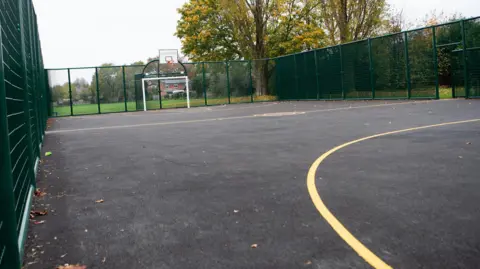 Charnwood Borough Council A multi-use games area with a grey surface, yellow lines and a football goal with a basketball net above at the end. The area is enclosed by green wire fencing.