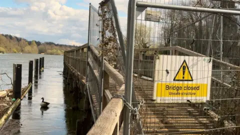 A footbridge fenced off with metal barriers. A duck is on the river.