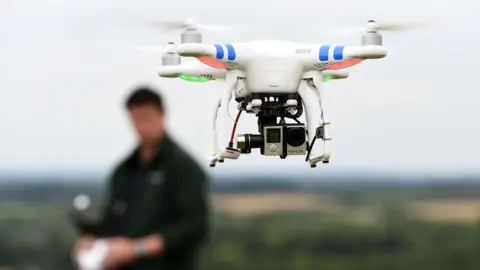 A drone in flight with the operator out of focus in the background.
