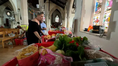 A woman with blonde hair and a black dress looking at a long table with fruit and vegetables on it inside a church with white painted walls