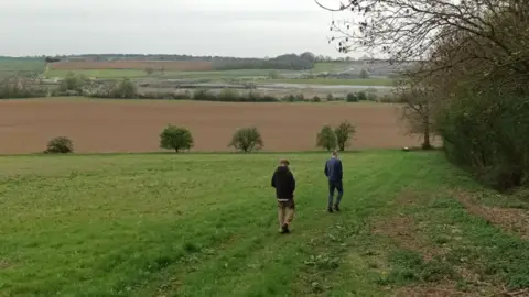 Northamptonshire Battlefields Society The back view of two men walking on the edge of a green pasture, with trees on their right. Ahead of them is a ploughed field and beyond that fields and hedges including a site where earth used to be.