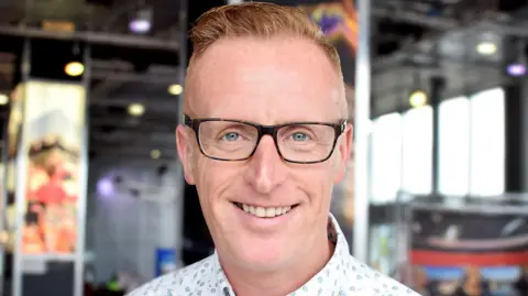Bryan Burnett, a man with short orange hair which is slightly spiked up. He is wearing black glasses and a patterned shirt.
