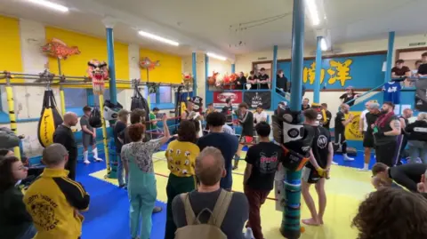 A group of spectators are in the gym at Hove Methodist Church. Some watch from balconies whilst others are ringside, as two fighters spar within it.