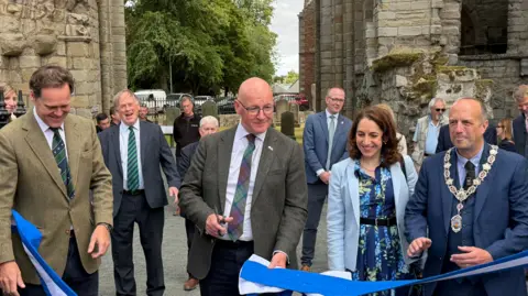 A line-up of people behind a blue and white ribbon which has been cut by Scotland's First Minister John Swinney